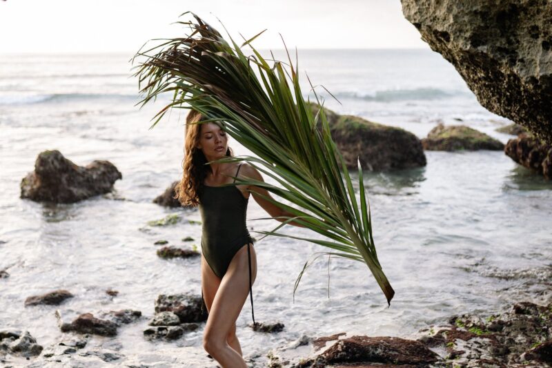 young woman with palm branch walking on rocky coastline